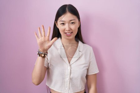 Foto de Mujer joven china de pie sobre fondo rosa mostrando y señalando hacia arriba con los dedos número cinco mientras sonríe confiado y feliz. - Imagen libre de derechos
