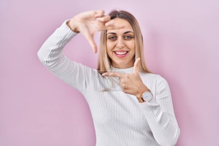Téléchargez les photos : Jeune femme blonde portant un pull blanc sur fond rose souriant faisant cadre avec les mains et les doigts avec le visage heureux. concept de créativité et photographie. - en image libre de droit