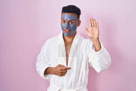 Téléchargez les photos : Young hispanic man wearing beauty face mask and bath robe waiving saying hello happy and smiling, friendly welcome gesture - en image libre de droit