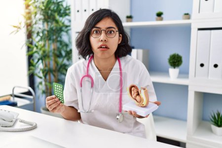 Photo for Young hispanic doctor woman holding anatomical model of uterus with fetus and birth control pills in shock face, looking skeptical and sarcastic, surprised with open mouth - Royalty Free Image