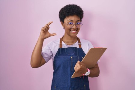 Photo for Young african american woman wearing professional waitress apron holding clipboard smiling and confident gesturing with hand doing small size sign with fingers looking and the camera. measure concept. - Royalty Free Image