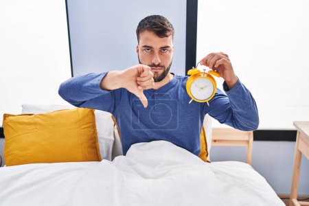 Foto de Handsome hispanic man in the bed holding alarm clock with angry face, negative sign showing dislike with thumbs down, rejection concept - Imagen libre de derechos
