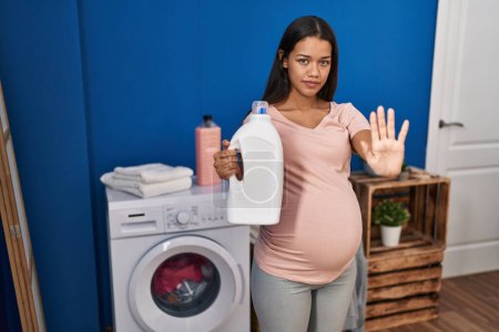 Téléchargez les photos : Jeune femme enceinte faisant la lessive tenant bouteille de détergent avec la main ouverte faisant stop signe avec expression sérieuse et confiante, geste de défense - en image libre de droit