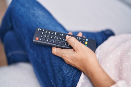 Foto de Mujer de pelo gris de mediana edad sosteniendo el control remoto de la televisión sentada en el sofá en casa - Imagen libre de derechos