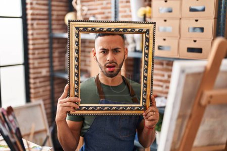 Photo for Young hispanic man sitting at art studio with empty frame clueless and confused expression. doubt concept. - Royalty Free Image