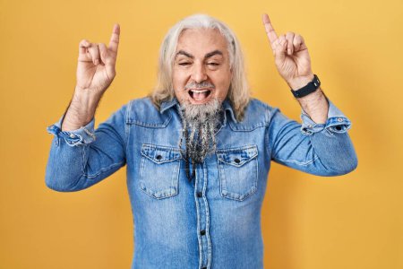 Foto de Middle age man with grey hair standing over yellow background smiling amazed and surprised and pointing up with fingers and raised arms. - Imagen libre de derechos