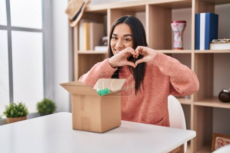 Photo for Young south asian woman opening cardboard box smiling in love doing heart symbol shape with hands. romantic concept. - Royalty Free Image