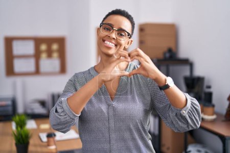 Téléchargez les photos : Femme afro-américaine travaillant au bureau portant des lunettes souriant dans l'amour faisant forme de symbole de coeur avec les mains. concept romantique. - en image libre de droit