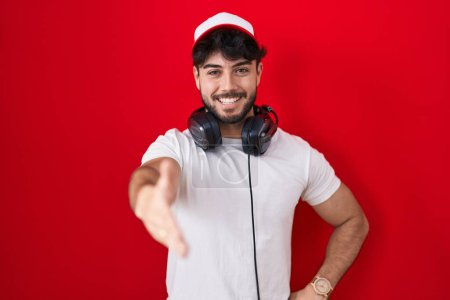 Foto de Hombre hispano con barba con sombrero de gamer y auriculares sonriendo amigable ofreciendo apretón de manos como saludo y bienvenida. negocios exitosos. - Imagen libre de derechos