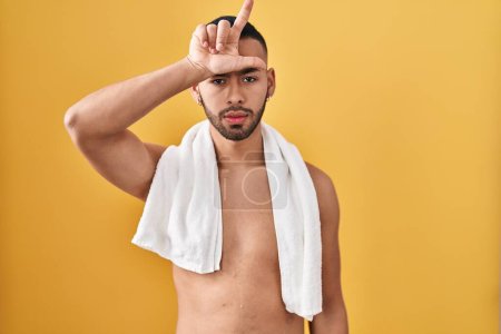 Téléchargez les photos : Young hispanic man standing shirtless with towel making fun of people with fingers on forehead doing loser gesture mocking and insulting. - en image libre de droit