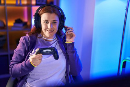 Foto de Young beautiful plus size woman streamer playing video game using joystick at gaming room - Imagen libre de derechos