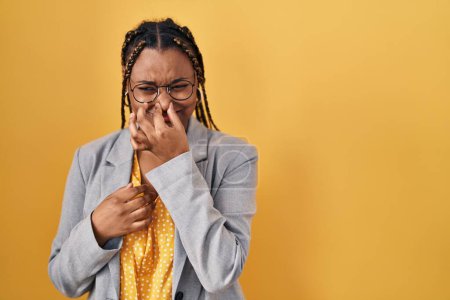 Téléchargez les photos : Femme afro-américaine avec des tresses debout sur fond jaune sentant quelque chose de puant et dégoûtant, odeur intolérable, retenant souffle avec les doigts sur le nez. mauvaise odeur - en image libre de droit
