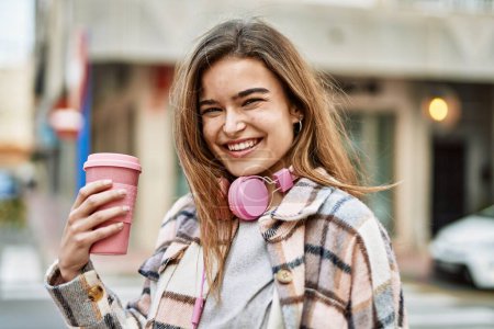 Téléchargez les photos : Jeune femme blonde portant des écouteurs tenant du café dans la rue - en image libre de droit