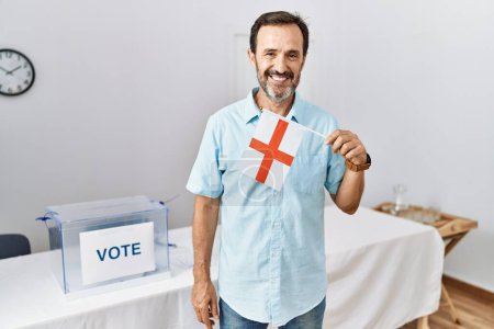 Téléchargez les photos : Homme d'âge moyen avec la barbe à l'élection de campagne politique tenant drapeau anglais regardant positif et heureux debout et souriant avec un sourire confiant montrant les dents - en image libre de droit