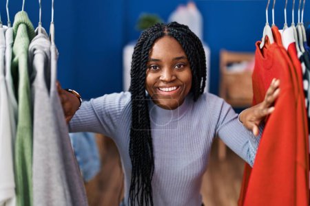 Téléchargez les photos : Femme afro-américaine à la recherche de vêtements sur porte-vêtements souriant avec un sourire heureux et cool sur le visage. montrant les dents. - en image libre de droit