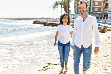 Foto de Middle age hispanic couple smiling happy walking with hands together at the beach. - Imagen libre de derechos
