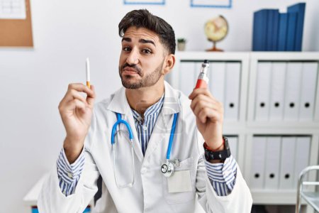 Photo for Young doctor man holding electronic cigarette at medical clinic smiling looking to the side and staring away thinking. - Royalty Free Image