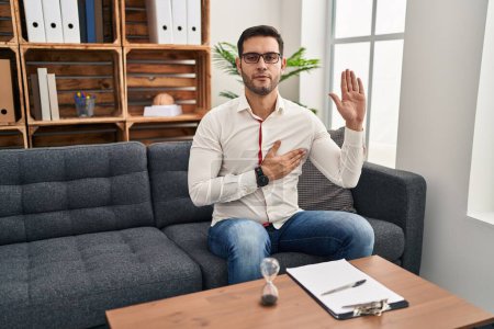 Téléchargez les photos : Jeune homme hispanique avec barbe travaillant au bureau de consultation jurant avec la main sur la poitrine et la paume ouverte, faisant un serment de fidélité - en image libre de droit