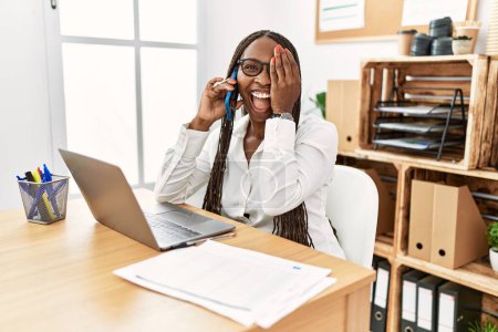 Foto de Mujer negra con trenzas trabajando en la oficina hablando por teléfono cubriendo un ojo con la mano, sonrisa confiada en la cara y emoción sorpresa. - Imagen libre de derechos