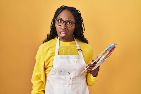 Téléchargez les photos : Femme africaine tenant la palette peintre en état de choc visage, l'air sceptique et sarcastique, surpris avec la bouche ouverte - en image libre de droit