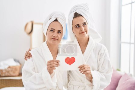 Téléchargez les photos : Femme d'âge moyen et fille portant une robe de bain tenant la carte de coeur souriant regardant vers le côté et regardant loin en pensant. - en image libre de droit
