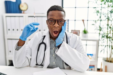 Téléchargez les photos : Jeune médecin africain tenant une seringue à l'hôpital effrayé et choqué, surprise et expression stupéfaite avec les mains sur le visage - en image libre de droit
