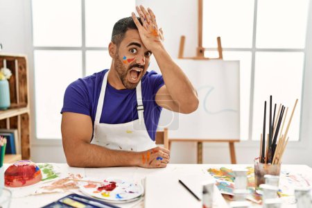 Téléchargez les photos : Jeune homme hispanique avec barbe au studio d'art avec visage peint surpris avec la main sur la tête pour erreur, rappelez-vous erreur. oublié, mauvais concept de mémoire. - en image libre de droit