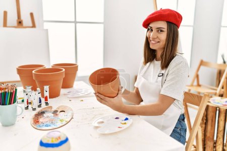 Photo for Young hispanic artist woman wearing french beret holding clay pot at art studio. - Royalty Free Image