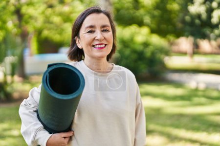 Foto de Mujer hispana de mediana edad sosteniendo esterilla de yoga en el parque luciendo positiva y feliz de pie y sonriendo con una sonrisa confiada mostrando los dientes - Imagen libre de derechos