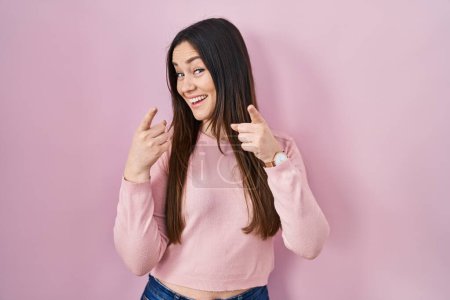 Téléchargez les photos : Jeune femme brune debout sur fond rose pointant les doigts vers la caméra avec un visage heureux et drôle. bonne énergie et vibes. - en image libre de droit