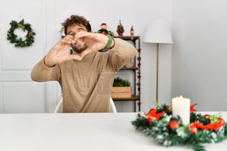 Téléchargez les photos : Jeune homme beau avec la barbe assis sur la table par la décoration de Noël souriant dans l'amour faisant forme de symbole de coeur avec les mains. concept romantique. - en image libre de droit