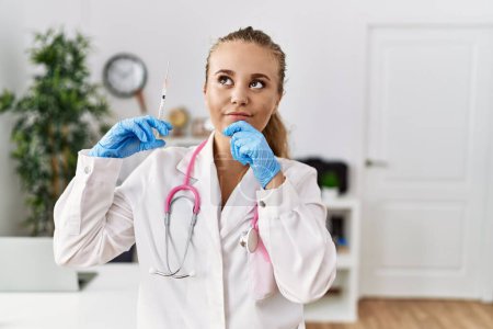 Photo for Young doctor woman holding syringe at the clinic serious face thinking about question with hand on chin, thoughtful about confusing idea - Royalty Free Image