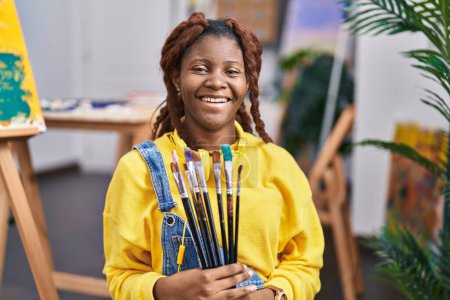 Foto de African american woman artist smiling confident holding paintbrushes at art studio - Imagen libre de derechos