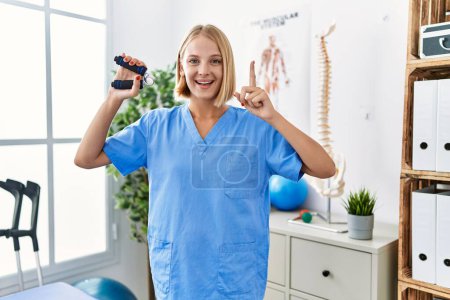 Téléchargez les photos : Young caucasian physiotherapist woman holding hand grip to train muscle smiling with an idea or question pointing finger with happy face, number one - en image libre de droit