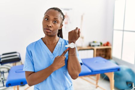 Téléchargez les photos : Femme noire avec des tresses travaillant à la clinique de récupération de la douleur à la hâte pointant vers regarder le temps, l'impatience, regardant la caméra avec une expression détendue - en image libre de droit