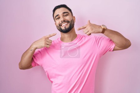 Foto de Joven hispano de pie sobre fondo rosa sonriendo alegre mostrando y señalando con los dedos los dientes y la boca. concepto de salud dental. - Imagen libre de derechos