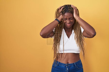 Foto de Mujer afroamericana con cabello trenzado de pie sobre fondo amarillo que sufre de dolor de cabeza desesperada y estresada porque el dolor y la migraña. manos en la cabeza. - Imagen libre de derechos