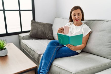 Foto de Mujer hispana de mediana edad sonriendo sentada en el sofá leyendo un libro en casa - Imagen libre de derechos