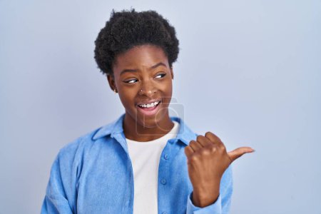 Foto de Mujer afroamericana de pie sobre fondo azul sonriendo con la cara feliz mirando y señalando a un lado con el pulgar hacia arriba. - Imagen libre de derechos