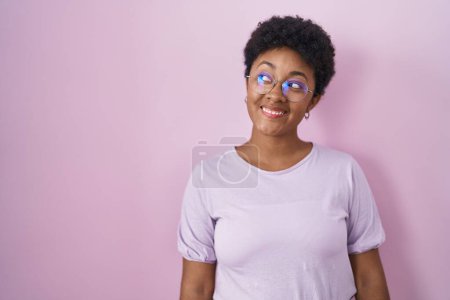 Foto de Joven mujer afroamericana de pie sobre fondo rosa sonriendo mirando hacia un lado y mirando hacia otro pensando. - Imagen libre de derechos