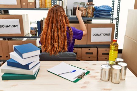 Téléchargez les photos : Jeune femme rousse portant un t-shirt volontaire sur le stand des dons posant vers l'avant avec la main du doigt - en image libre de droit