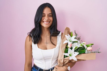 Foto de Mujer morena sosteniendo ramo de flores blancas guiñando el ojo mirando a la cámara con expresión sexy, cara alegre y feliz. - Imagen libre de derechos