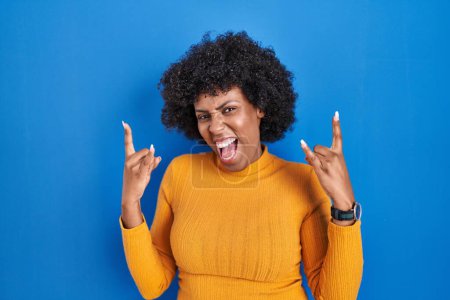 Photo for Black woman with curly hair standing over blue background shouting with crazy expression doing rock symbol with hands up. music star. heavy music concept. - Royalty Free Image