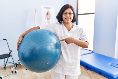 Téléchargez les photos : Jeune physiothérapeute hispanique femme tenant pilates balle souriant heureux pointant avec la main et le doigt - en image libre de droit