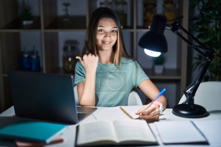 Téléchargez les photos : Adolescente faisant ses devoirs à la maison tard dans la nuit pointant le dos derrière avec la main et les pouces levés, souriant confiant - en image libre de droit