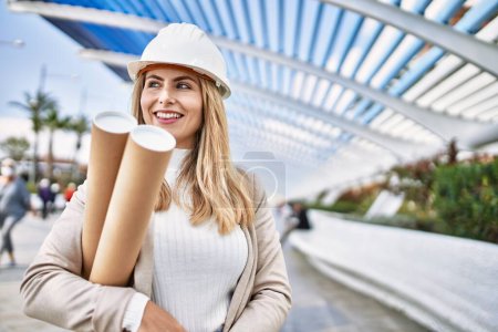 Photo for Young blonde woman wearing hardhat holding blueprints at street - Royalty Free Image