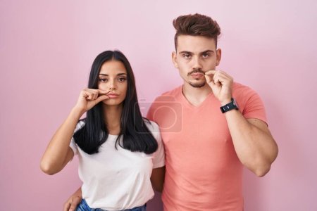 Junges hispanisches Paar, das über rosa Hintergrundmund steht und die Lippen als Reißverschluss mit den Fingern geschlossen hält. Geheimniskrämerei und Schweigen, Tabuthema 
