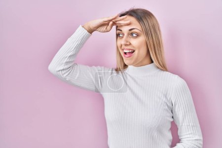 Photo for Young blonde woman wearing white sweater over pink background very happy and smiling looking far away with hand over head. searching concept. - Royalty Free Image