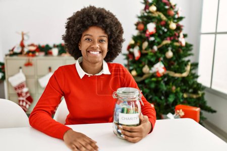 Foto de Joven mujer afroamericana sosteniendo tarro de caridad sentado junto al árbol de Navidad en casa - Imagen libre de derechos