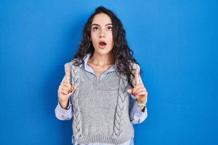 Photo for Young brunette woman standing over blue background amazed and surprised looking up and pointing with fingers and raised arms. - Royalty Free Image